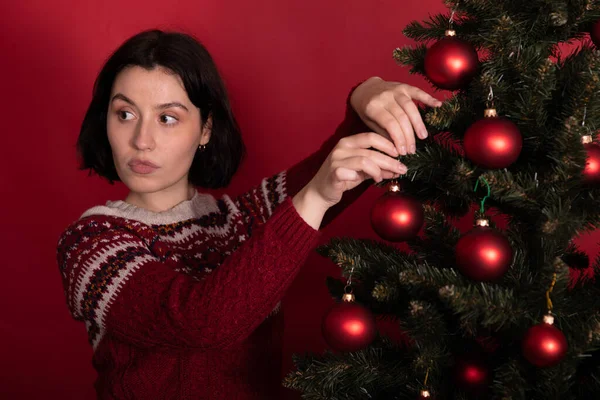 Omtänksam Brunett flicka hängande leksak bollar på julgran på röd bakgrund — Stockfoto
