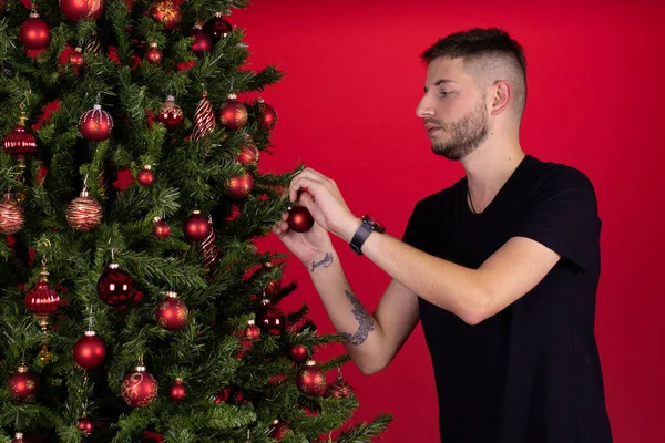 Joven hombre atractivo decorando árbol de Navidad sobre fondo rojo. Navidad sola — Foto de Stock