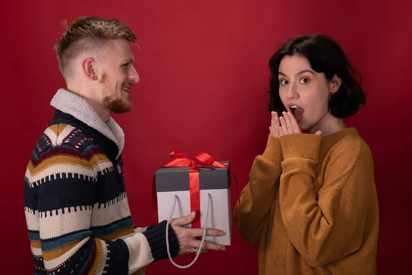 Feliz hombre presenta la caja de regalo de Navidad a la novia sorprendida sobre fondo rojo — Foto de Stock