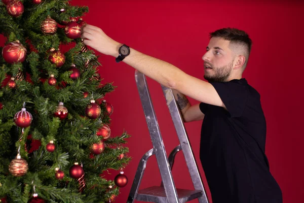 Unshaven man in black T-shirt hanging toy balls on Christmas tree using ladder