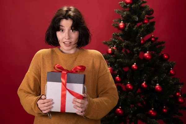 Jong mooi vrouw kijkt verrast houdt xmas cadeau op kerstboom achtergrond — Stockfoto