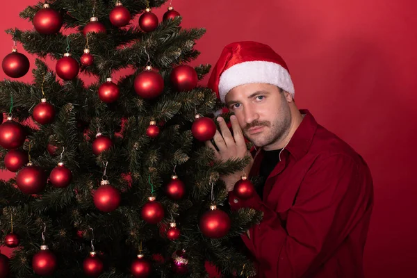 Hombre joven en el sombrero de Santa suavemente abraza el árbol de Navidad sobre fondo rojo, espacio de copia — Foto de Stock