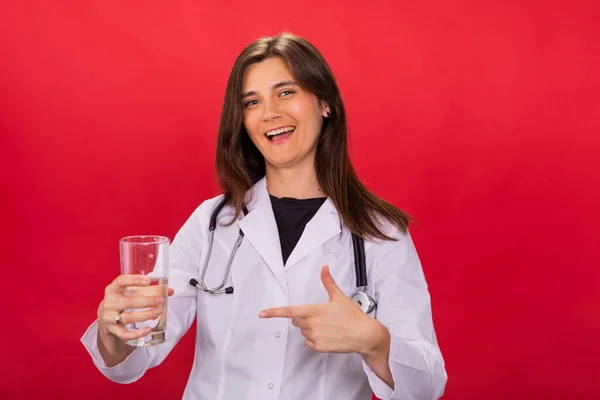 Attraktive brünette Ärztin zeigt mit dem Finger auf Glas mit klarem Wasser auf Rot — Stockfoto