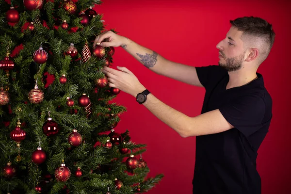 Joven tatuado sin afeitar en camiseta negra colgando pelotas de juguete en el árbol de Navidad — Foto de Stock