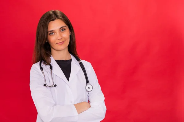 Jolie brunette femme médecin posant les bras croisés isolé sur rouge studio — Photo