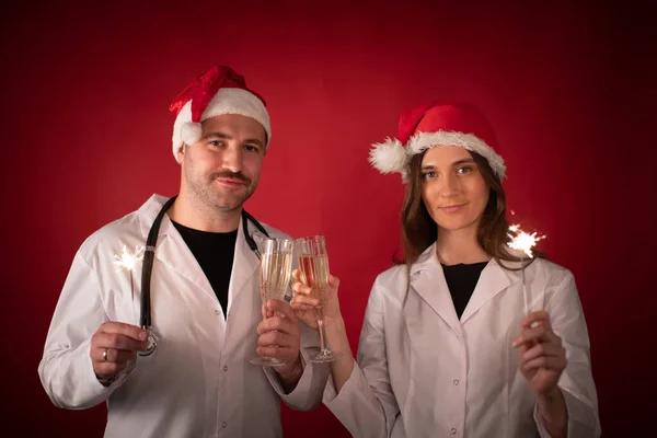 Två leende läkare i jultomten hattar hejar med champagne flöjter på rött — Stockfoto