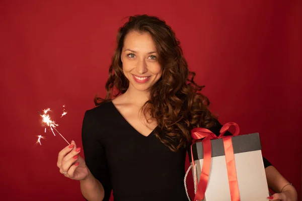 Encantadora dama con regalos navideños y chispa ardiente sonriendo sobre fondo rojo — Foto de Stock