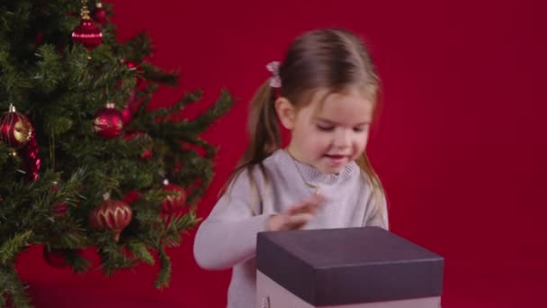 Piccola ragazza eccitata apertura scatola regalo di Natale vicino all'albero di Natale su sfondo rosso — Video Stock