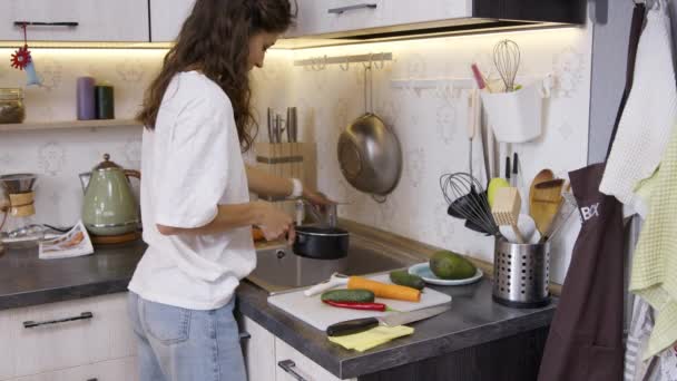 若い女性料理ビーガンディナーに自宅,大人と鍋に水を注ぐ — ストック動画