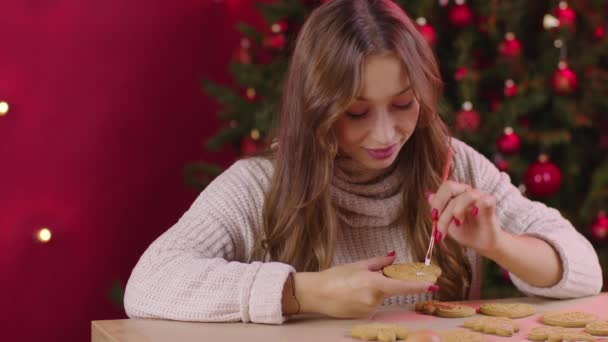 Bonita menina sorridente de cabelos compridos gelando biscoito de gengibre de Natal em forma de coração — Vídeo de Stock