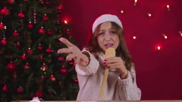 Fröhliche attraktive Frau mit Weihnachtsmann-Hut singt Weihnachtslieder mit Küchenspachtel — Stockvideo