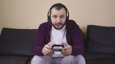 Kulaklıklı genç yakışıklı erkek oyuncu elinde joystick ile online video oyunu oynuyor.