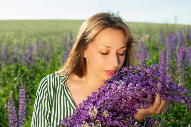 Güzel kız çiçek açmış tarlada oturuyor gözleri kapalı ve Lupin çiçeklerini kokluyor.