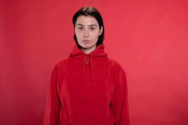 Brunette meisje in rood hoodie kijken naar camera eend lippen op rode studio achtergrond — Stockfoto