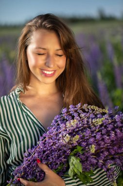 Mutlu gülümseyen kız güneş ışığında bir avuç kır çiçeğiyle gözlerini kısıyor.