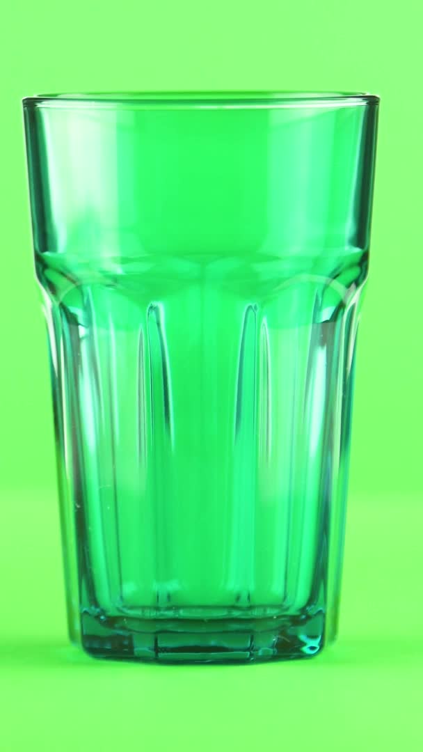 Вертикальное видео Сок наливается в зеленые граненые стекла изолированы на светло-зеленом фоне — стоковое видео
