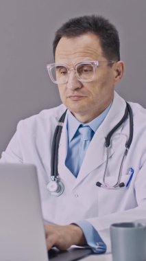 Dikey çekim. Tıp üniformalı yorgun doktor bilgisayarla çalışıyor. Bir yudum kahve alıyor.