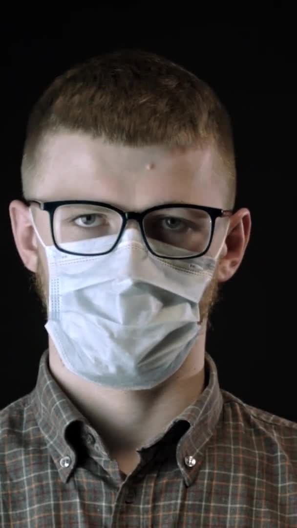 Vertikální záběr Mladý muž v lékařské masce proti covid-19 se podívá do kamery. Ohnisko koronaviru — Stock video