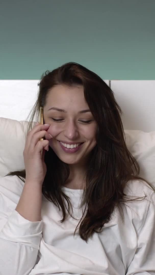 Tiro vertical feliz jovem mulher em pijama branco falando no celular na cama e sorrindo — Vídeo de Stock