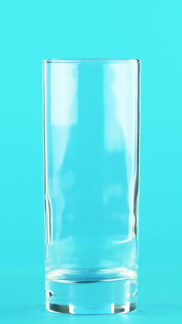 垂直视频4K特写镜头拍摄的橙汁冷饮倒入玻璃杯蓝色背景的工作室 — 图库视频影像