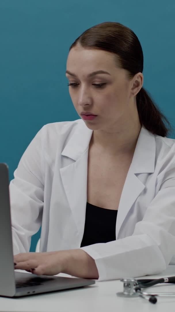 Vertikalt bilde Sexy kvinnelig lege i hvit legefrakk sitter ved et bord og arbeider på laptop – stockvideo