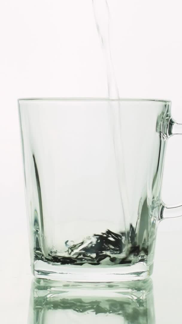 Vídeo vertical partículas de chá preto derramando em caneca transparente de vidro e adicionando água fervente para preparar chá isolado no fundo branco fechar — Vídeo de Stock