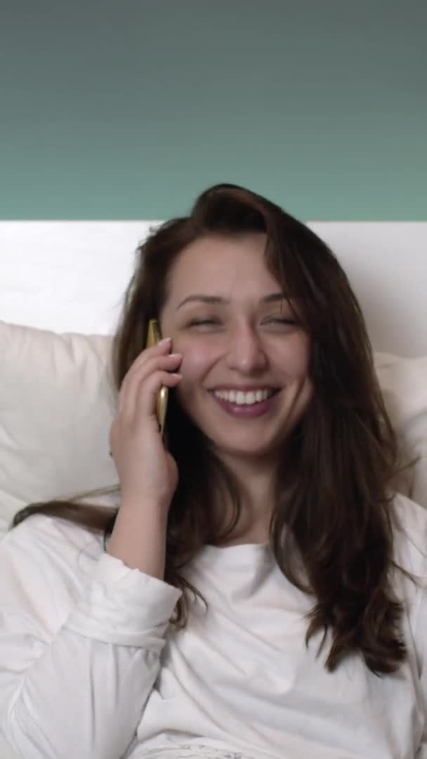 Κάθετη βολή Χαρούμενη καυκάσιος γυναίκα μιλάει στο κινητό τηλέφωνο στο κρεβάτι και χαμογελά γενικά — Αρχείο Βίντεο