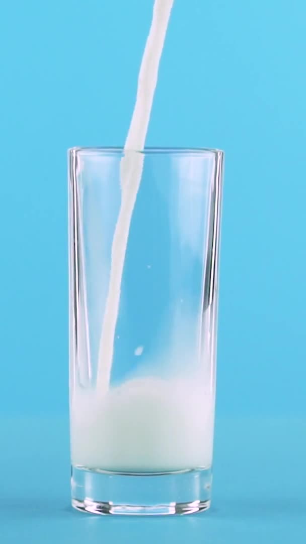 Vídeo vertical Slow motion close-up shot of milk cold beverage pooring into low glass blue background in studio — Vídeo de Stock