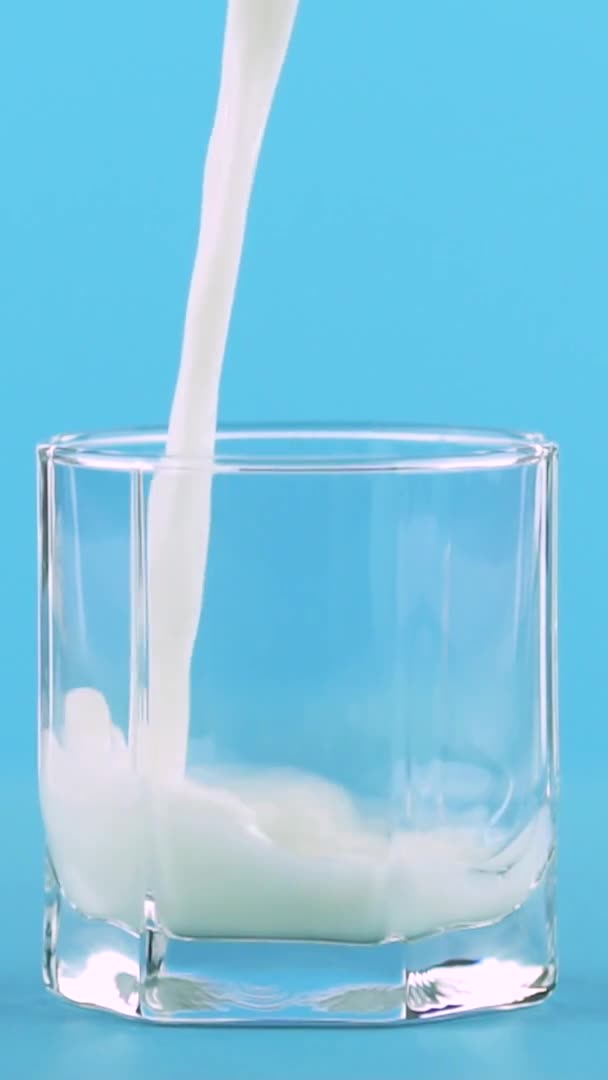 Vídeo vertical Primer plano en cámara lenta de la bebida de leche fría pooring en vidrio bajo con facetas de fondo azul en el estudio — Vídeos de Stock