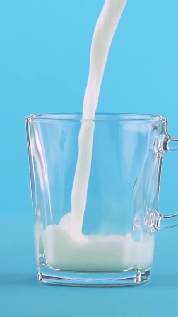 Vídeo vertical Primer plano en cámara lenta de la bebida de leche fría pooring bebida en taza de vidrio con mango fondo azul en el estudio — Vídeos de Stock
