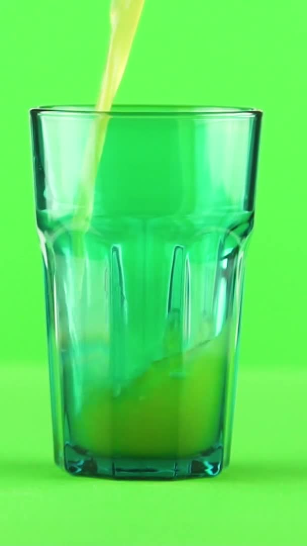 Вертикальное видео Сок наливается в зеленое огранённое стекло изолированное на светло-зеленом фоне замедленного движения — стоковое видео
