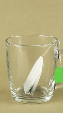 Yeşil etiketli dikey siyah çay poşeti şeffaf bardağa konur ve bej arka planda izole edilmiş kaynar suyla doldurulur.