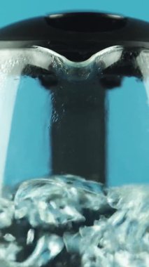 Dikey video. Şeffaf duvarları olan elektrikli su ısıtıcısı. Kaynama işlemi. Mavi arkaplan