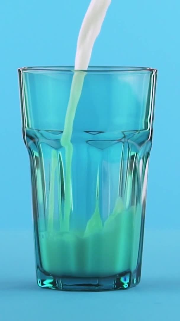 Vídeo vertical Primer plano en cámara lenta de la bebida de leche fría pooring en fondo azul de cristal facetado azul en el estudio — Vídeos de Stock