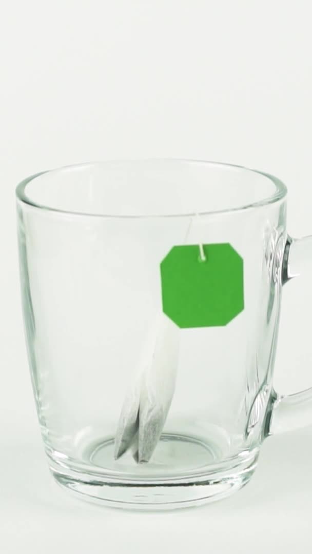 Vertikale Video schwarzer Teebeutel mit grünem Etikett wird in einem transparenten Becher aus Glas platziert und mit kochendem Wasser gefüllt, isoliert auf weißem Hintergrund in Zeitlupe — Stockvideo