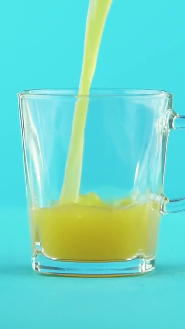 Vídeo vertical Primer plano en cámara lenta de zumo de fruta naranja multifruta bebida fría pooring en taza de vidrio con mango fondo azul en estudio — Vídeos de Stock