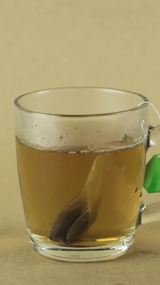 垂直ビデオ黒ティーバッグ持ち上げとガラス透明なマグカップに落下ベージュの背景スローモーションで隔離されたお茶を醸造する — ストック動画