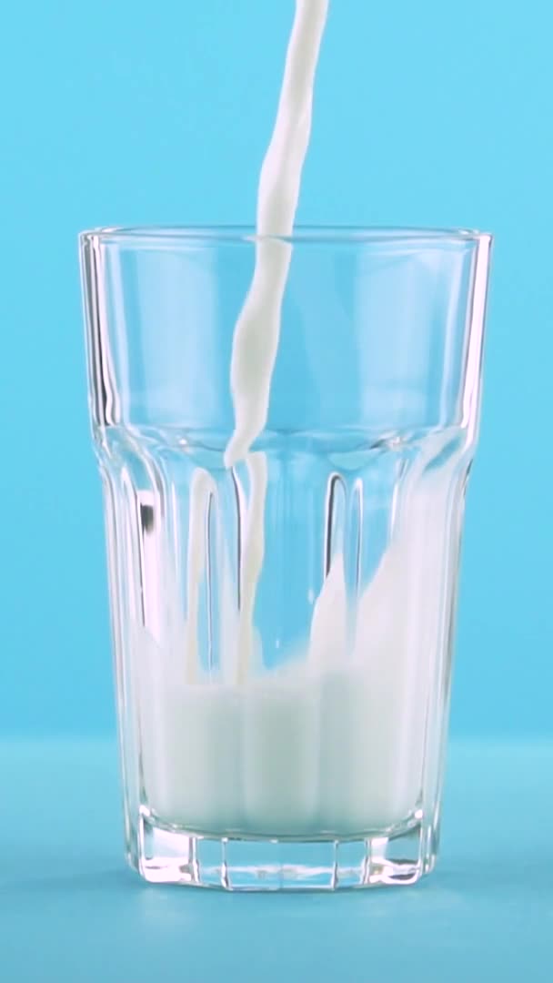 Vídeo vertical Primer plano en cámara lenta de la bebida de leche fría pooring bebida en vidrio facetado sobre fondo azul de color en el estudio — Vídeos de Stock