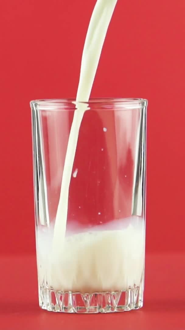 Вертикальное видео Медленное движение крупным планом снимок холодного молочного напитка холодного дневника, переливающегося в резьбовое стекло красного фона в студии — стоковое видео