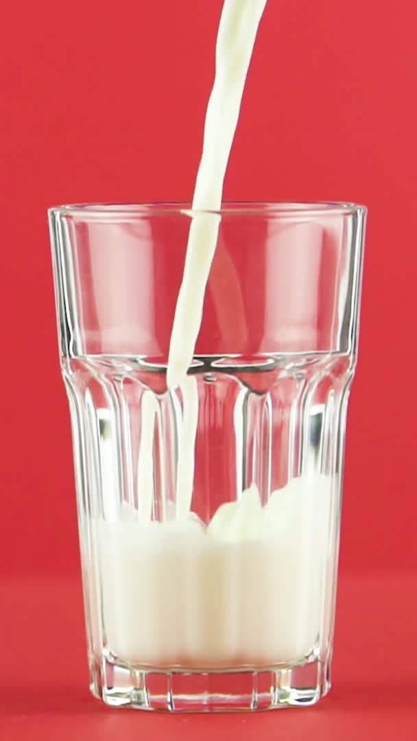 Vídeo vertical Slow motion close-up shot of cold diary milk cold beverage pooring into faceted glass on colored red background in studio — Vídeo de Stock