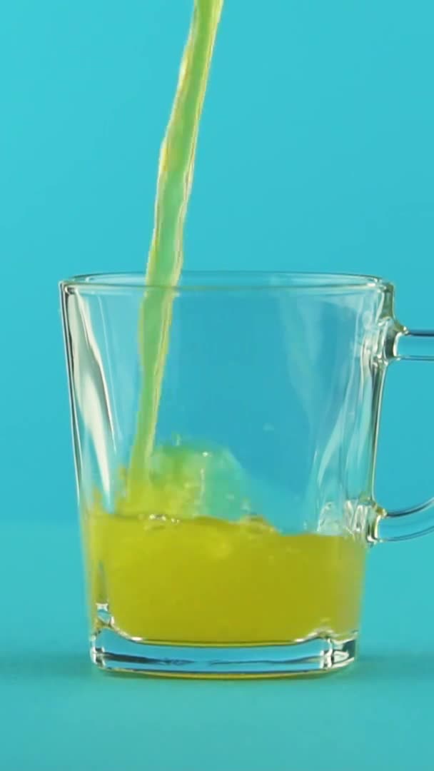 Függőleges videó Lassú mozgás közeli felvétel gyümölcsös szénsavas narancsos üdítő hideg ital pooring üvegbögrébe kék háttér a stúdióban — Stock videók