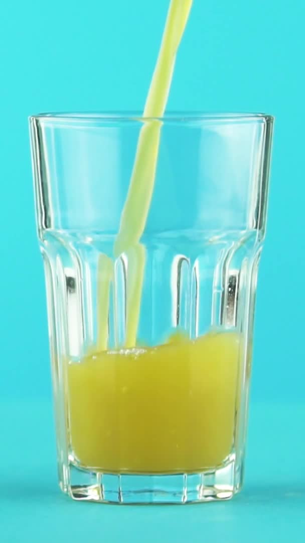 Vídeo vertical Primer plano en cámara lenta de zumo de fruta naranja multifruta bebida fría pooring en vidrio facetado sobre fondo azul de color en el estudio — Vídeos de Stock