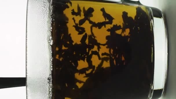 Вертикальная видеоложка перемешивает чай в прозрачной стеклянной чашке, создавая вихрь, черные частицы, вращающиеся пивоваренный чай. замедленное движение на белом фоне вблизи — стоковое видео