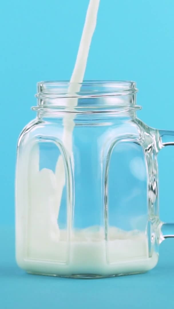 Vídeo vertical Primer plano en cámara lenta de la bebida de leche fría pooring en frasco de vidrio fondo azul en el estudio — Vídeo de stock