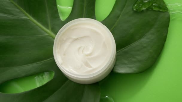 Tarro cosmético con crema sobre hoja tropical verde en salpicadura de agua limpia, vista superior — Vídeos de Stock