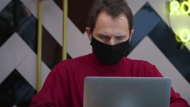 Knappe man van middelbare leeftijd in zwart beschermend masker werken met laptop in cafe — Stockvideo