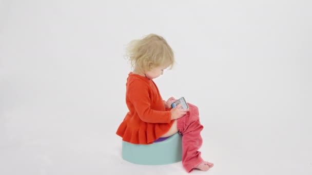 Rozczochrane dziecko siada na nocniku dla dzieci za pomocą smartfona na białym tle — Wideo stockowe