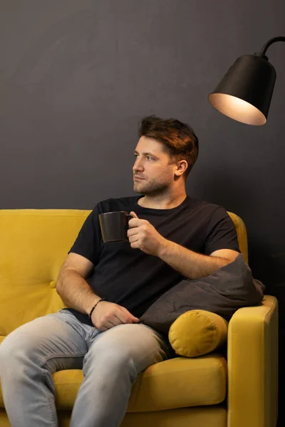 Hombre aburrido sin afeitar con taza de café mirando hacia otro lado sentado en cómodo sofá amarillo — Foto de Stock