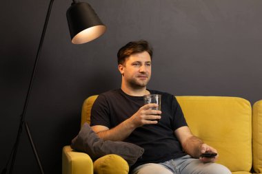 Genç traşsız adam kanepede içki ve uzaktan kumandayla film izliyor.