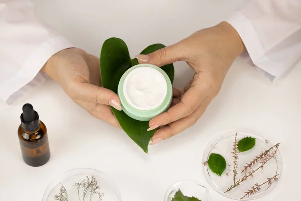 Hands hold cosmetic cream jar on green leaf on white lab background, dermatology Royalty Free Stock Images